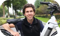 A man in casual attire smiles while kneeling next to a bicycle in a park, with a green pathway lined with trees in the background.