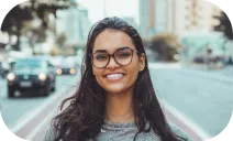 A photo of a smiling woman with long dark hair and glasses, standing on a city street with buildings and cars in the background.