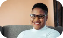 This is a photo of a smiling person with short hair, wearing glasses, hoop earrings, and a white top, seated against a beige background.
