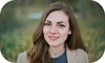 A close-up photograph of a young woman with long brown hair, light makeup, and a slight smile, set against a blurred outdoor background.