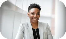 This is a color photograph of a smiling person wearing a light gray blazer and black shirt, standing indoors against a light background.