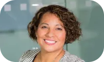 A woman with short curly hair is smiling in front of a blurred office background.