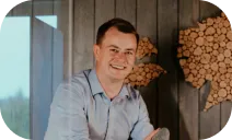 A man wearing a light blue shirt is smiling in front of a wooden wall with wooden circular art pieces.