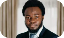 A professional portrait of a man with a black beard and afro hairstyle, dressed in a black suit, white shirt, and tie.