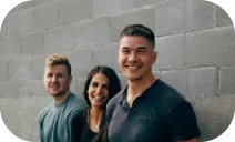 This is a photo of three smiling people standing side by side in front of a gray brick wall.